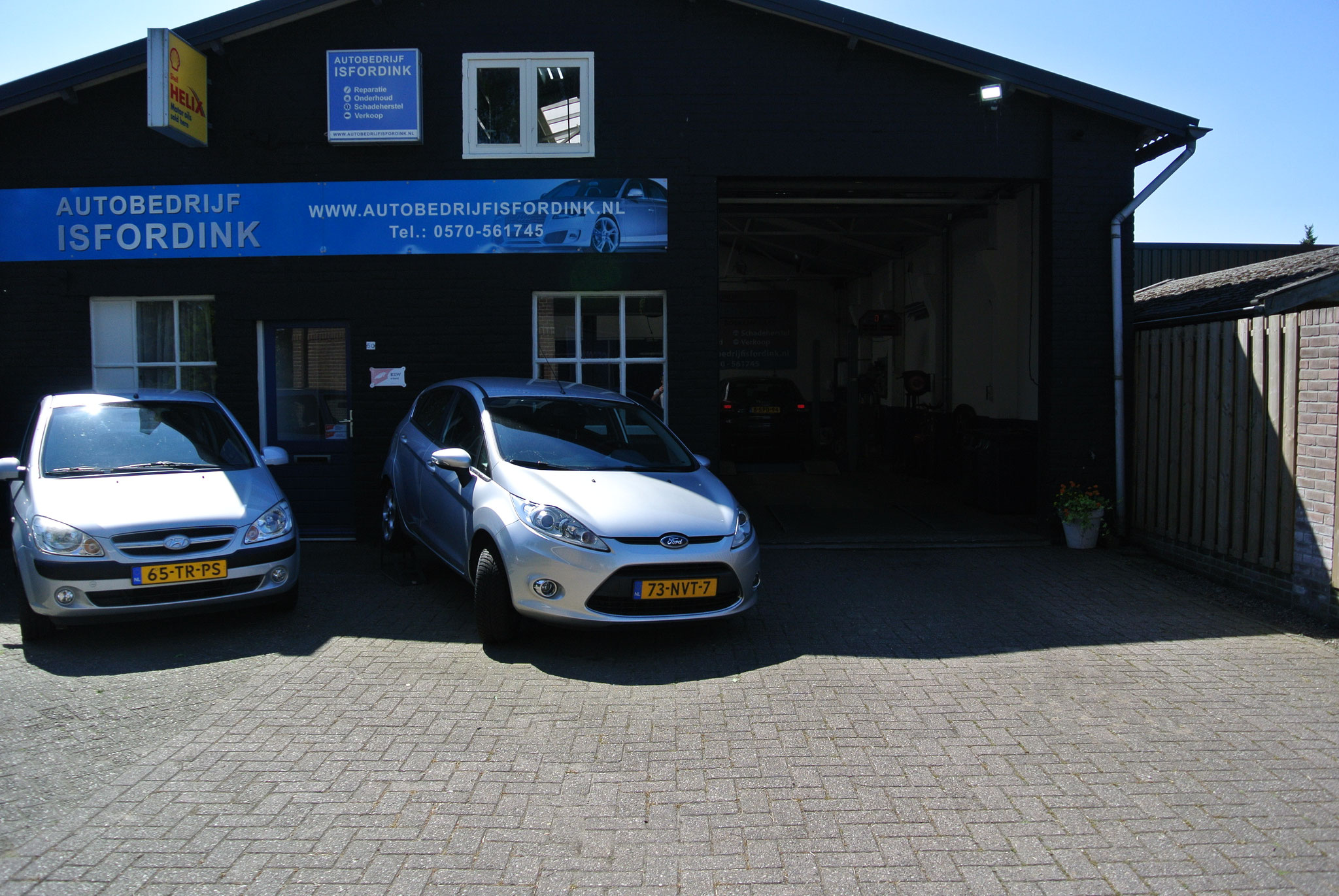 De voorkant van het pand met de open werkplaatsdeur en twee auto's ervoor